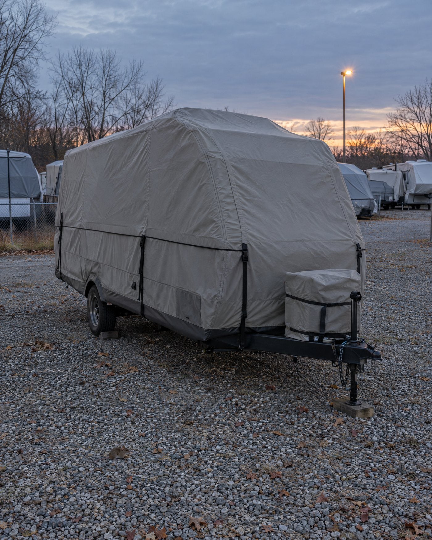 A covered travel trailer parked in a gravel RV storage lot at dusk